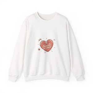 May include: White sweatshirt with a graphic of a broken heart in shades of red and pink. The heart has the text "Love Stinks, Let's Drink!" in a playful font. Wine glasses and confetti surround the heart.