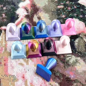 以下が含まれることがあります： ピンク、青、紫、虹色の様々な色合いのスタンプを含む、カラフルなダイヤモンドペインティングツールのコレクション。ツールは模様のある表面に配置され、1つのスタンプが分離されています。