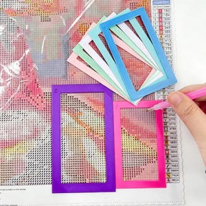 Può includere: Accessori colorati per la pittura con diamanti. L'immagine mostra cornici rettangolari viola, rosa, blu e verdi. Una mano tiene uno stilo rosa ed è visibile un dipinto con diamanti parzialmente completato.