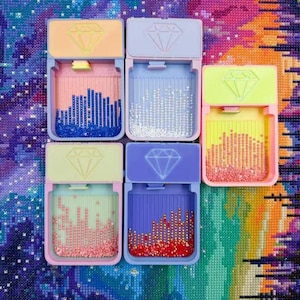 May include: Six colorful diamond painting organizers with hinged lids. Each organizer is a different pastel color, with a diamond graphic on the lid. The containers hold various colors of diamond painting drills, creating a vibrant display.
