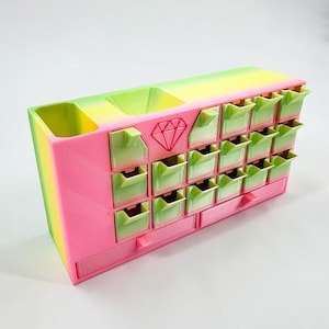 May include: A multi-compartment organizer in a gradient of pink, yellow, and green. It features small, square drawers and larger open compartments. A red diamond graphic is on the front. This storage unit is ideal for small items.