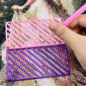 May include: A pink and purple diamond painting tray with a grid pattern. A pink stylus is held over the tray, with a diamond being placed. The tray is designed to hold and organize small craft items for diamond painting.