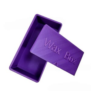 Op de afbeelding: Een paarse rechthoekige waxdoos met een deksel. Het deksel staat iets open en onthult de binnenkant van de doos. De woorden "Wax Box" zijn in reliëf op het deksel aangebracht. De doos is gemaakt van een stevig materiaal.