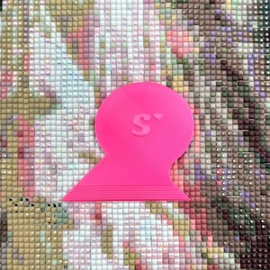 Könnte beinhalten: Ein leuchtend rosa Diamant-Malwerkzeug mit abgerundeter Oberseite und einer flachen, geriffelten Kante. Der Buchstabe "S" ist oben sichtbar. Das Werkzeug befindet sich vor dem Hintergrund eines unfertigen Diamantgemäldes.