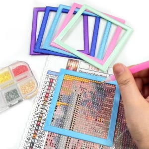 Puede incluir: Accesorios para pintura de diamantes. La imagen muestra una variedad de marcos cuadrados de colores, un pequeño recipiente con cuentas de colores y un proyecto de pintura de diamantes en progreso. Una mano sostiene un lápiz óptico rosa.