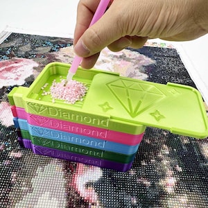May include: A stack of colourful diamond painting trays in green, pink, blue, and purple. Each tray is labelled "Diamond". A hand uses a pink stylus to pick up pink diamond-shaped beads from the green tray.