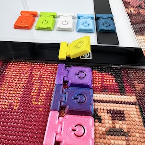 Könnte beinhalten: Eine Sammlung farbenfroher Lichtpad-Clips in Orange, Grün, Weiß, Hellblau und Dunkelblau. Die Clips sind auf einem Lichtpad angeordnet, mit zusätzlichen Clips in Gelb, Lila und Rosa. Jeder Clip zeigt ein Symbol für die Ein-/Aus-Taste.