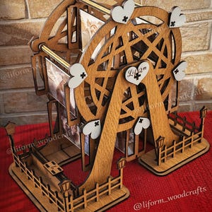 Puede incluir: Una exhibición de fotos de rueda de la fortuna de madera con detalles de corazón. El diseño incluye una valla decorativa y pequeñas linternas. Se ven las palabras "DESCARGA DIGITAL" y "ARCHIVOS DE CORTE LÁSER". El diseño es de madera.
