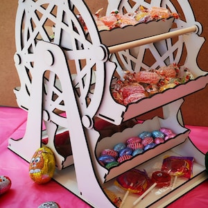 Può includere: Un espositore di caramelle a forma di ruota panoramica in legno bianco, con tre livelli di vassoi pieni di caramelle incartate e lecca lecca. L'espositore è decorato con dettagli neri e tasselli di legno. Uova di Pasqua al cioccolato sulla superficie rosa.