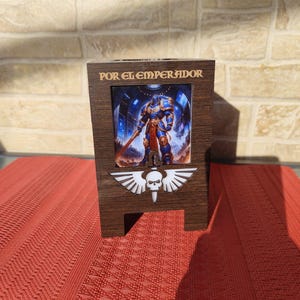 May include: A wooden display stand featuring a framed image of a futuristic warrior in blue armor, with the text "POR EL EMPERADOR" at the top. The stand has a white skull and wing emblem. The stand is on a red surface.