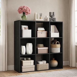 May include: A black cube storage unit with nine compartments, filled with decorative items like vases, boxes, and a bouquet of pink flowers. The unit is placed against a neutral-colored wall, with a window and curtains visible.