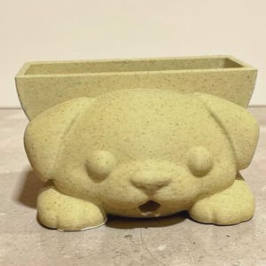 May include: A light yellow dog-shaped planter with a rectangular opening on top. The planter has a textured surface and a cute, cartoonish design. The dog's face features large eyes, a small nose, and a slightly open mouth. The planter is approximately 6 inches long.