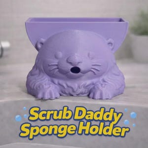 Op de afbeelding: Een paarse Scrub Daddy spons houder in de vorm van een otter. De houder heeft een rechthoekig compartiment voor een spons en een schattig otter gezicht. De tekst "Scrub Daddy Sponge Holder" staat eronder.