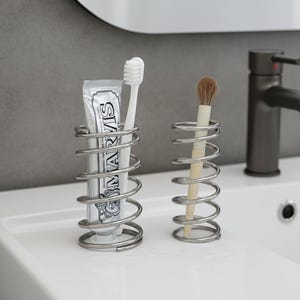 May include: Two spiral-shaped metal holders on a white surface. One holds a tube of toothpaste and a white toothbrush. The other holds a makeup brush. The toothpaste tube reads "MARVIS".