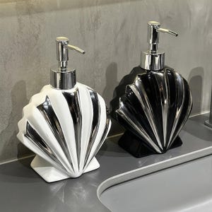 May include: Two seashell-shaped soap dispensers. One is white with silver accents, the other is black with silver accents. Both have a silver pump top. The dispensers are on a grey countertop, suitable for a bathroom.