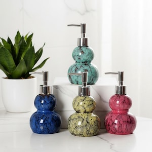 Può includere: Quattro dispenser di sapone in ceramica a forma di doppia zucca con finitura smaltata crepata. I dispenser sono color turchese, blu scuro, giallo e rosa, ciascuno sormontato da un meccanismo a pompa in metallo argentato per il liquido.