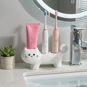 May include: A white ceramic cat-shaped toothbrush holder with two electric toothbrushes, one white and one pink. A pink tube of toothpaste and a small potted succulent are also visible. The holder has a whimsical design.