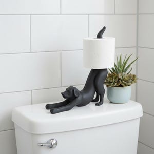 May include: A black dog-shaped toilet paper holder. The dog is in a stretching pose, supporting a roll of white toilet paper. A small potted succulent sits beside the holder on a white toilet.