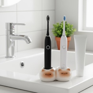 May include: Two electric toothbrushes, one black and one white, are in a double-sided ceramic holder with cat faces. A white tube of toothpaste sits to the right of the toothbrushes on a white sink. A chrome tap is in the background.