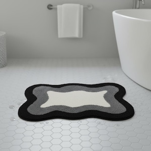 Puede incluir: Una alfombra de baño con una forma única y un diseño de color en capas. La alfombra presenta un borde negro, una capa gris medio, una capa gris claro y un centro blanco. La alfombra está sobre un suelo de baldosas blancas.