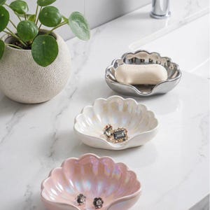 Peut inclure: Trois coupelles en forme de coquillage, déclinées en argenté, blanc irisé et rose irisé. La coupelle argentée contient un savon, tandis que les coupelles blanche et rose contiennent des petites boucles d'oreilles. Finition brillante.