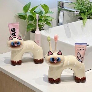 Puede incluir: Dos porta cepillos de dientes con forma de gato crema con patas y detalles marrones, ojos azules y orejas triangulares negras. Uno sostiene un tubo de pasta de dientes rosa y el otro cepillos de dientes.
