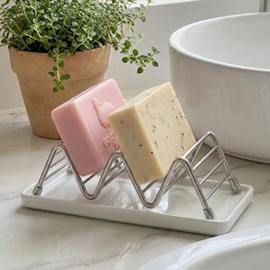Peut inclure: Un porte-savon en céramique blanche avec un support en zigzag en acier inoxydable. Deux savons reposent dans le support : une barre rose avec un motif de chèvre et une barre beige avec des taches sombres. Une plante en pot est en arrière-plan.