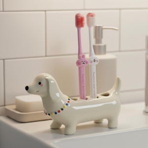 May include: A cream-coloured dachshund-shaped toothbrush holder with a colourful beaded collar. Two toothbrushes, one pink and one white, are inserted into the holder. The toothbrushes have cartoon animal designs.