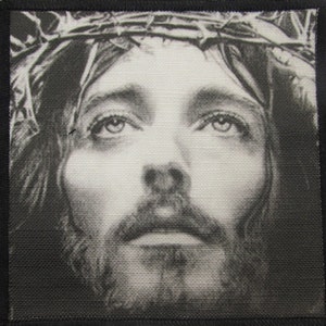 Può includere: Immagine in bianco e nero di un viso maschile con una corona di spine sulla testa. L'immagine è stampata su una toppa di tessuto.