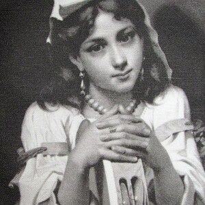 May include: A black and white portrait of a young girl wearing a white bonnet and a pearl necklace. She is holding a book in her hands and looking at the viewer.
