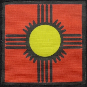 Peut inclure: Un carré rouge avec un symbole Zia noir au centre. Le symbole Zia est un soleil stylisé avec quatre rayons s'étendant du centre. Le symbole est un symbole traditionnel du peuple Pueblo du Nouveau-Mexique.