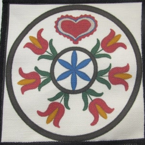 Op de afbeelding: Een wit vierkant van stof met een zwarte rand, met een cirkelvormig ontwerp met een rood hart, een blauwe bloem en zes rode tulpen met gele centra.