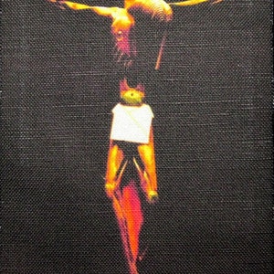 May include: A red and yellow image of a person on a cross. The person is depicted with a white cloth around their waist. The background is black.
