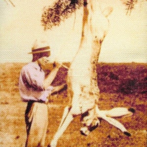 Könnte beinhalten: Ein Mann mit Hut und hellem Hemd steht neben einem Baum. Er hält ein Messer in der Hand und scheint ein Reh zu häuten.