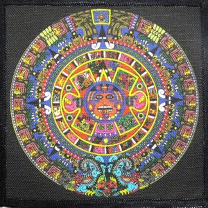 May include: A colorful circular patch with a detailed design of the Aztec calendar, featuring a central sun god with a red face and a large tongue. The calendar is surrounded by a ring of glyphs and symbols in various colors, including blue, yellow, red, and green.