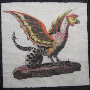 Op de afbeelding: Een vierkante patch met een mythisch wezen ontwerp. Het wezen heeft een hanenkop, vleugels in oranje, geel en groen en een gestreept lichaam. De patch is zwart omrand. Het wezen staat op een bruine rots.