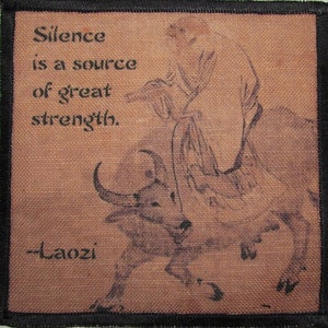 May include: A brown fabric patch with a black border featuring a black and white illustration of a person riding a bull. The text "Silence is a source of great strength. -Laozi" is printed on the patch.