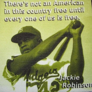 May include: A black and white photo of Jackie Robinson, a baseball player, wearing a baseball uniform with the words "Dodgers" and "Jackie Robinson" on it. The quote "There's not an American in this country free until every one of us is free." is printed above the photo.