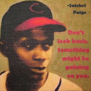May include: A black and red baseball cap with a white "C" on the front. The cap is worn by a person with dark skin. The text "Don't look back, something might be gaining on you." is written in pink on a tan background. The text "Satchel Paige" is written in black on the tan background.