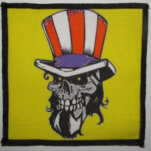 Puede incluir: Una calavera en blanco y negro que lleva un sombrero de copa rojo, blanco y azul con una banda morada. La calavera tiene una larga barba negra y está sobre un fondo amarillo.