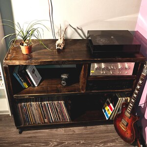 Könnte beinhalten: Ein dunkel gebeizter Holz-Medien-Schrank mit Plattenspieler, Verstärker und Schallplatten. Eine Gitarre lehnt an der Seite. Das obere Regal enthält eine kleine Topfpflanze, einen Schädel und Bücher. Der Schrank hat eine rustikale, Vintage-Ästhetik.