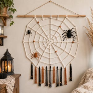 May include: A cream-colored macrame spider web wall hanging suspended from a wooden dowel. The web features small wooden beads, one large black crocheted spider, one small black spider, and one orange spider. Black and orange tassels hang below.