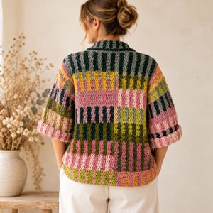 Pode incluir: Um cardigã de crochê com um padrão de listras verticais em cores alternadas de verde escuro, verde claro, amarelo mostarda e rosa suave. A peça possui gola, mangas três quartos com punhos dobrados e uma bainha reta.