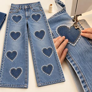 May include: Light blue denim jeans featuring multiple dark blue heart-shaped patches with frayed white edges. The jeans have a classic five-pocket design with a button and zipper fly closure. The patches are stitched onto the front of the legs.