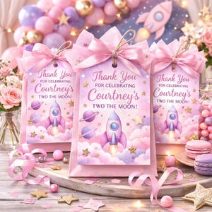 May include: Three pink gift bags with tags that read "Thank You for Celebrating Courtney's Two the Moon!" Each tag features a rocket ship, planets, and stars. The bags are tied with pink bows and are surrounded by party decorations.