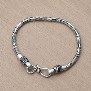 May include: A silver-toned snake chain bracelet featuring a textured, woven metal pattern. The bracelet is secured with a hook-and-eye clasp mechanism, which includes two decorative cylindrical end caps with intricate dotted and ribbed detailing.