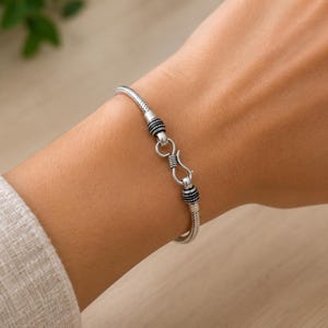 Op de afbeelding: Een zilverkleurige slangenkettingarmband met een getextureerde, flexibele metalen band. De armband wordt vastgezet met een opvallende S-haaksluiting, geflankeerd door twee kleine, donker geoxideerde decoratieve kralen met gedetailleerde stippen.