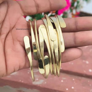 Puede incluir: Un brazalete de metal en tono dorado que presenta múltiples bandas delgadas y curvas superpuestas con extremos suaves y redondeados. El diseño consta de varias tiras pulidas interconectadas que crean una apariencia fluida. El texto bhawnakcjewellers es visible.