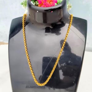 Puede incluir: Un collar de cadena de color dorado que presenta una serie de eslabones circulares uniformes y entrelazados. La cadena se muestra sobre un soporte negro, destacando el acabado metálico y el patrón consistente de los eslabones metálicos redondos.