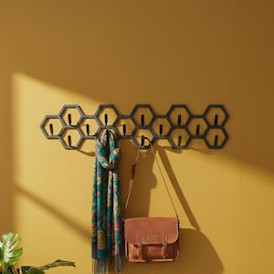 May include: A dark gray honeycomb-shaped wall-mounted coat rack with multiple hooks. A teal scarf and a brown leather satchel hang from the hooks. The rack is mounted on a yellow wall.